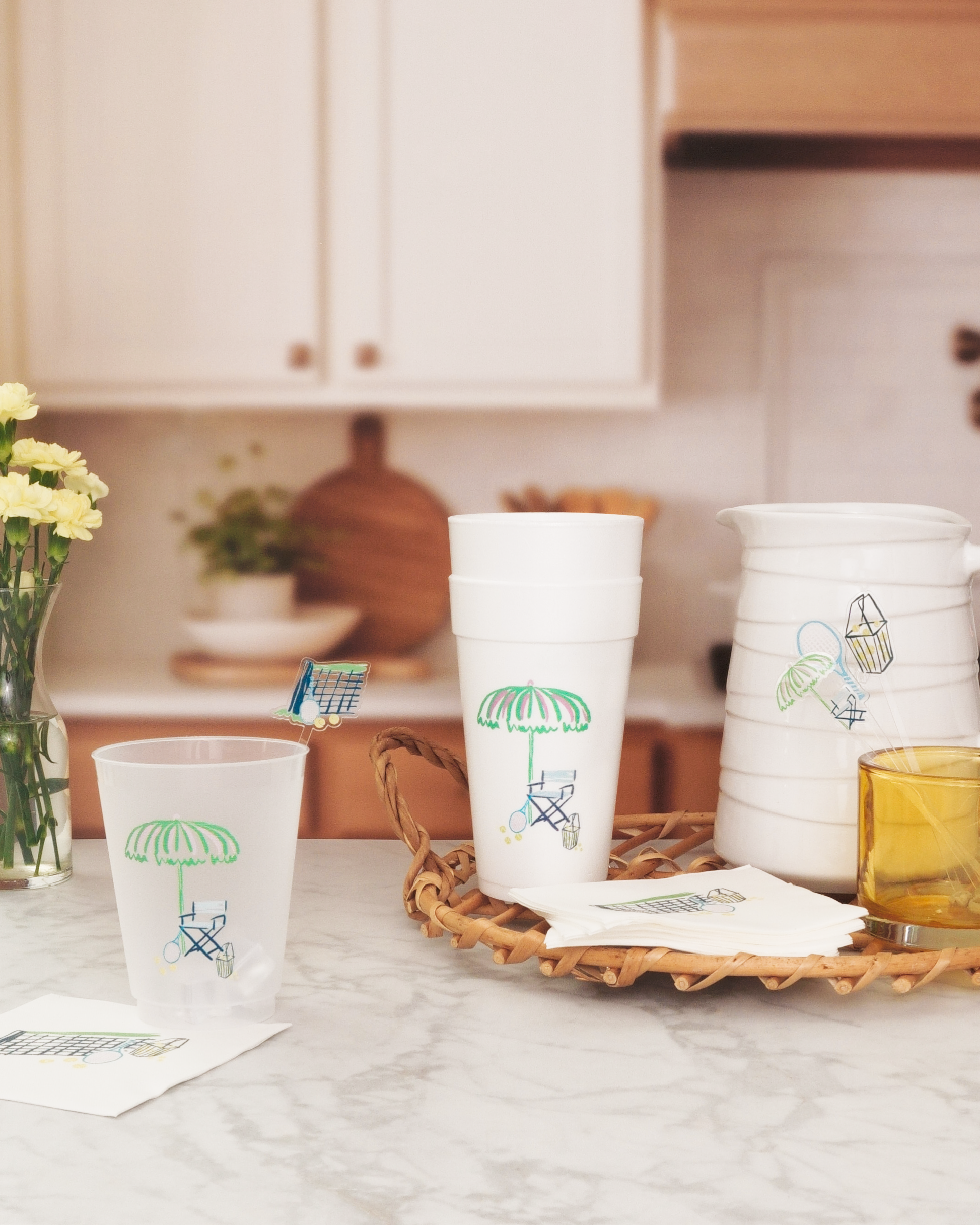 a kitchen counter with various items, including a white pitcher, a white cup with an umbrella design, and a basket. There are also some yellow flowers and a vase on the counter.