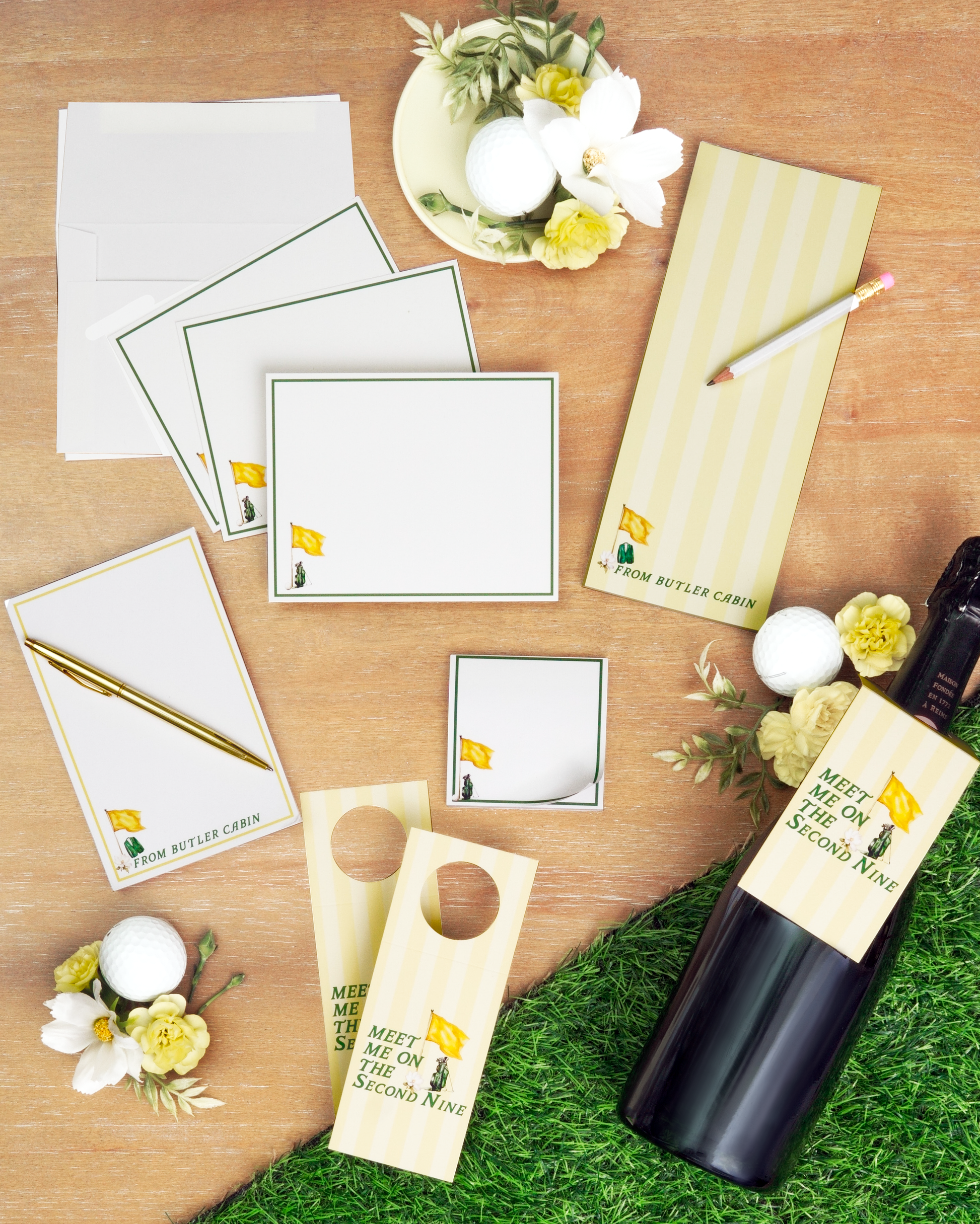 a collection of items related to golf, including a bottle of wine, a pen, a card, and a note pad, all arranged on a wooden surface with a green grass background.