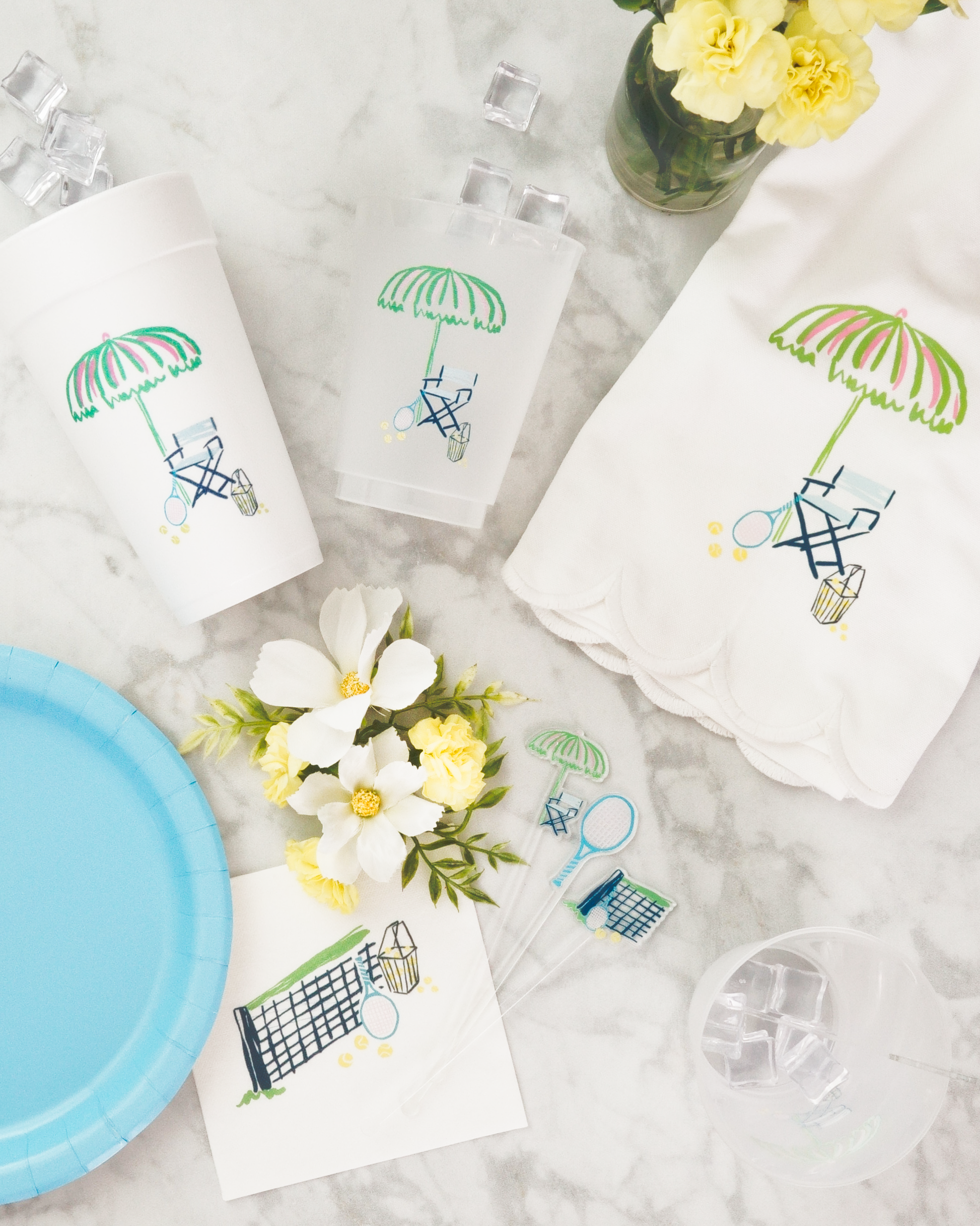 a collection of summer-themed items, including a white paper cup with a green umbrella design, a white napkin with a green umbrella design, a blue plate with a tennis racket and flowers, and a vase with yellow flowers.