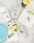 a collection of summer-themed items, including a white paper cup with a green umbrella design, a white napkin with a green umbrella design, a blue plate with a tennis racket and flowers, and a vase with yellow flowers.